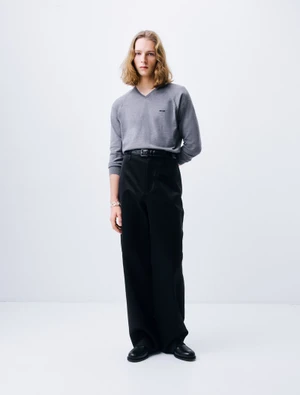 Model with long blonde hair wearing a grey v-neck sweater and black trousers against white seamless background