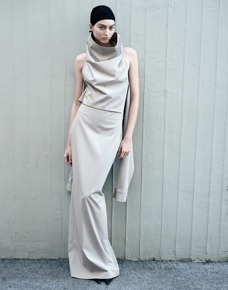 Model wearing a structured beige cowl-neck top and long skirt with a black cap against a vertical paneled wall