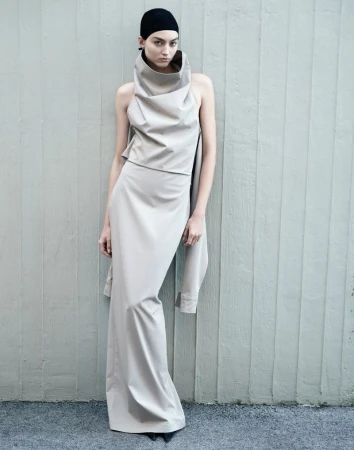 Model wearing a structured beige cowl-neck top and long skirt with a black cap against a vertical paneled wall