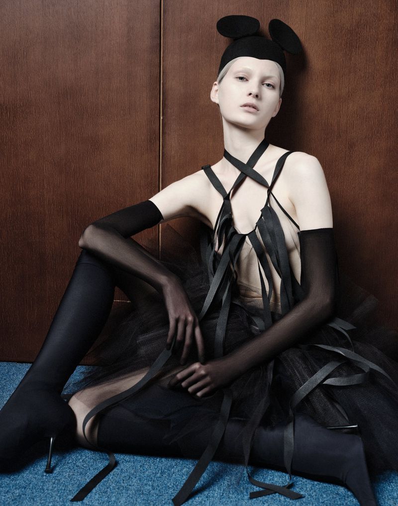 Model in black avant-garde outfit with mouse-ear cap sitting against a wood panel wall