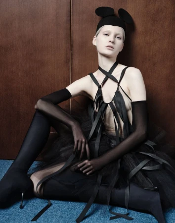 Model in black avant-garde outfit with mouse-ear cap sitting against a wood panel wall