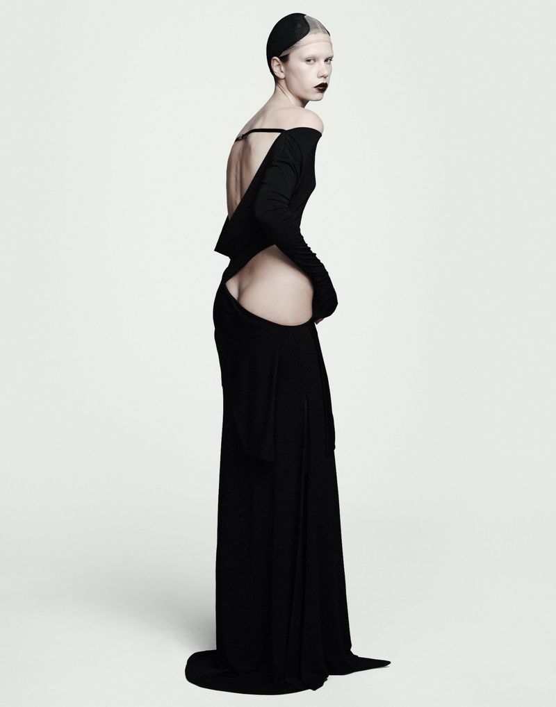 Model in a long black evening gown looking back over her shoulder against a plain white studio background.