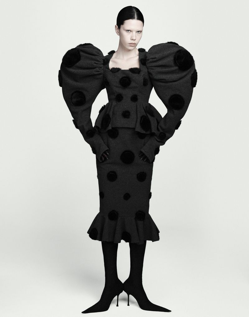 Model wearing a black polka dot dress with exaggerated puffy shoulders and heeled boots against a white studio background.