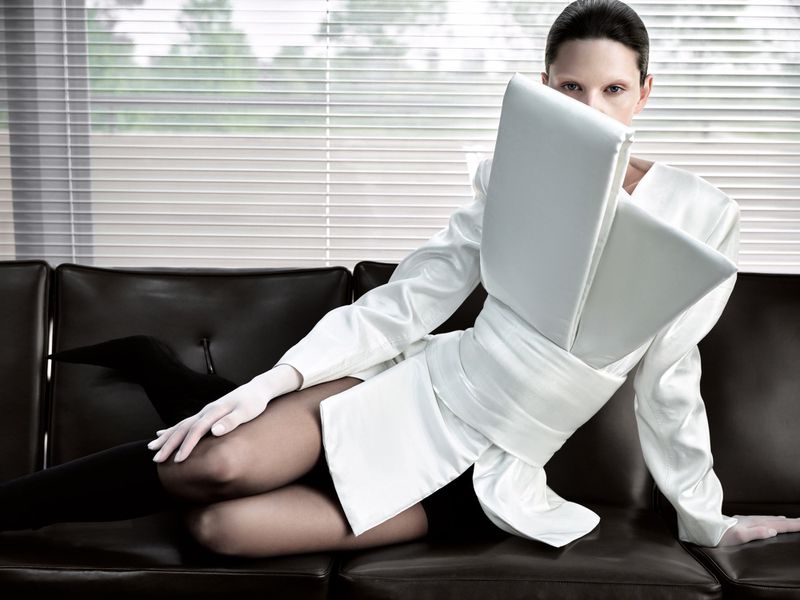 Model wearing sculptural white garment leaning on a dark leather couch in front of horizontal window blinds