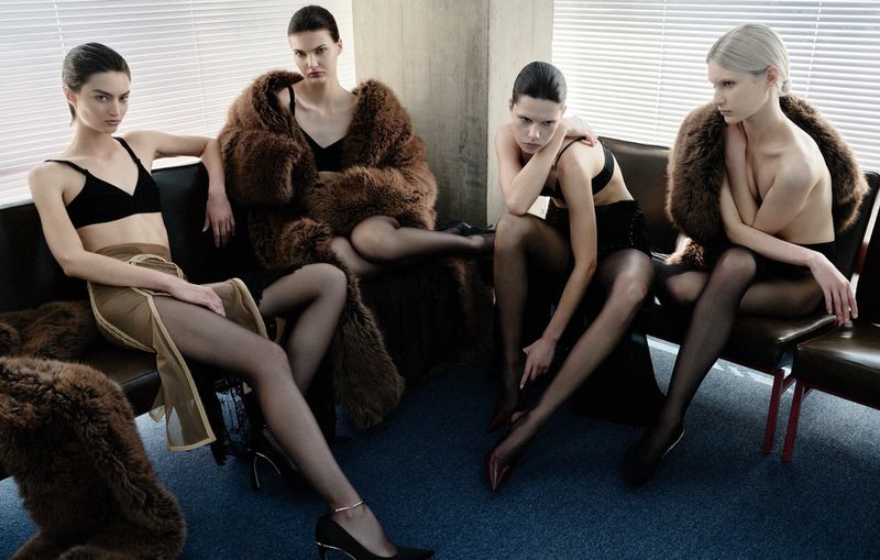 Four women in lingerie and coats sit on a bench in a bright indoor setting with minimalist style, creating a fashion or beauty editorial.