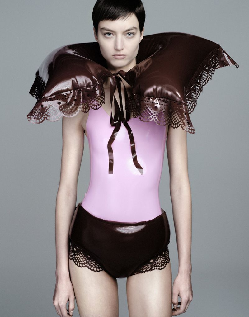 Model wearing a glossy brown puffed collar and matching bottoms with a pink bodysuit against a neutral grey studio background