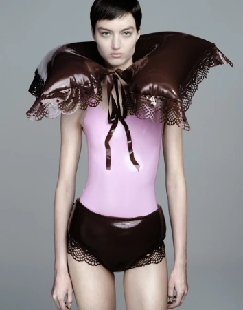 Model wearing a glossy brown puffed collar and matching bottoms with a pink bodysuit against a neutral grey studio background