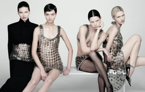 Four female models in embellished metallic and black garments posing together on a white bench against a light grey background.