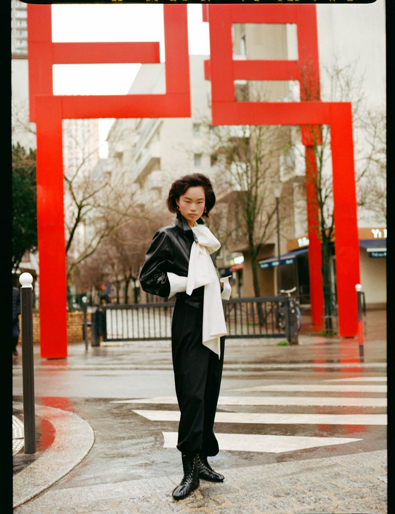 Model standing outdoors on a wet street wearing a black jacket and white scarf beneath a red structure