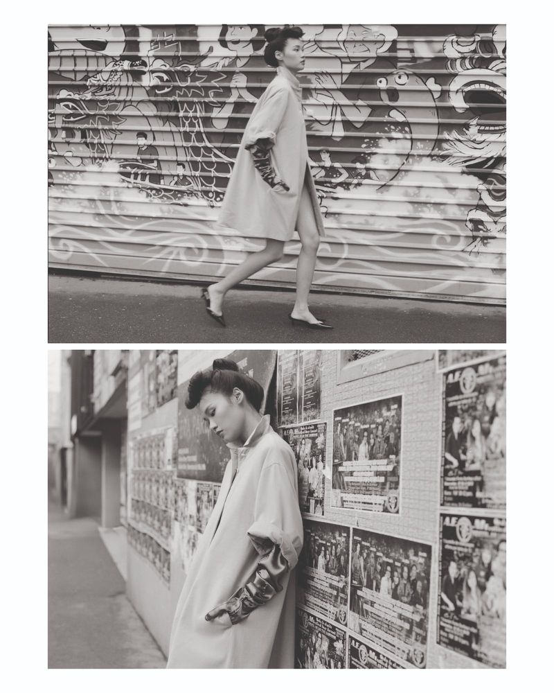 Model walking past a graphic roller shutter and leaning against a poster-covered alley wall in black and white