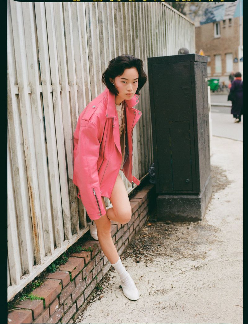 Model in a bright pink leather jacket leaning against a white wooden fence outdoors on a street.