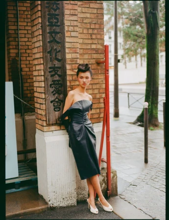 Model in strapless dark leather dress leaning against a brick pillar with a wooden sign on a city street.