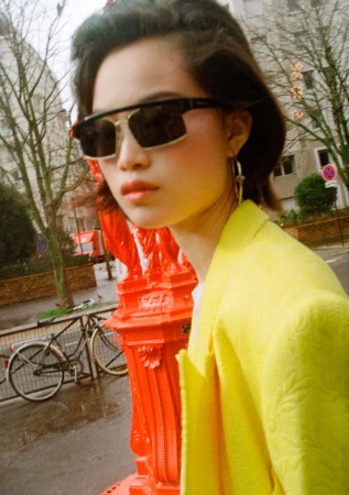 Close-up of a model wearing dark sunglasses and a bright yellow jacket outdoors by an urban street