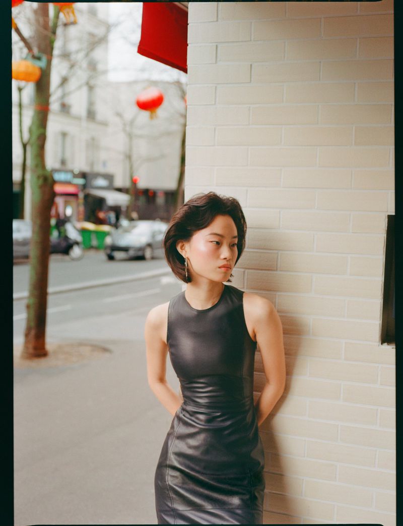 Model in a black sleeveless dress leaning against a cream brick wall on a city street with red lanterns