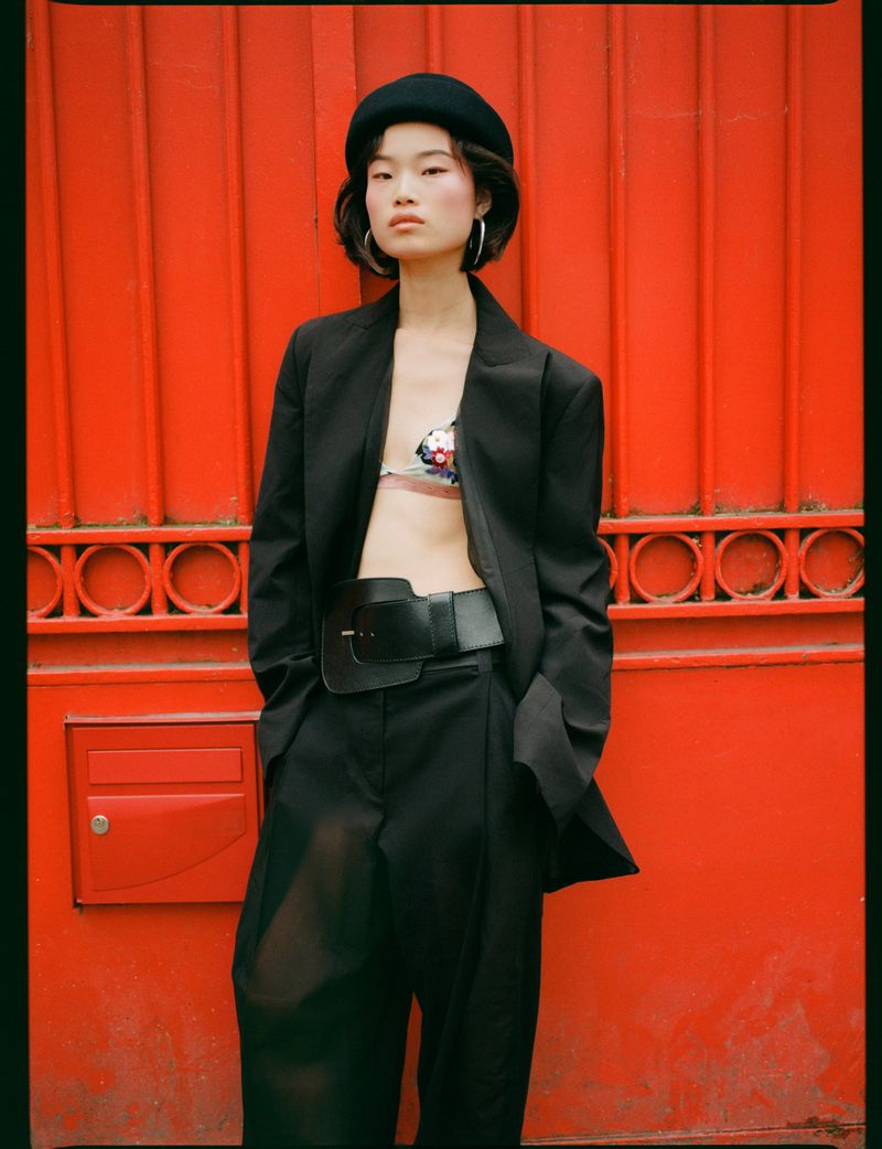 Model wearing a black blazer, matching trousers, wide belt, and beret against a vibrant red panel wall.