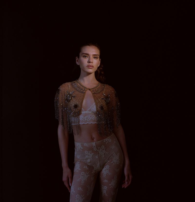 Portrait of a woman wearing a beaded cape and patterned trousers against a dark background