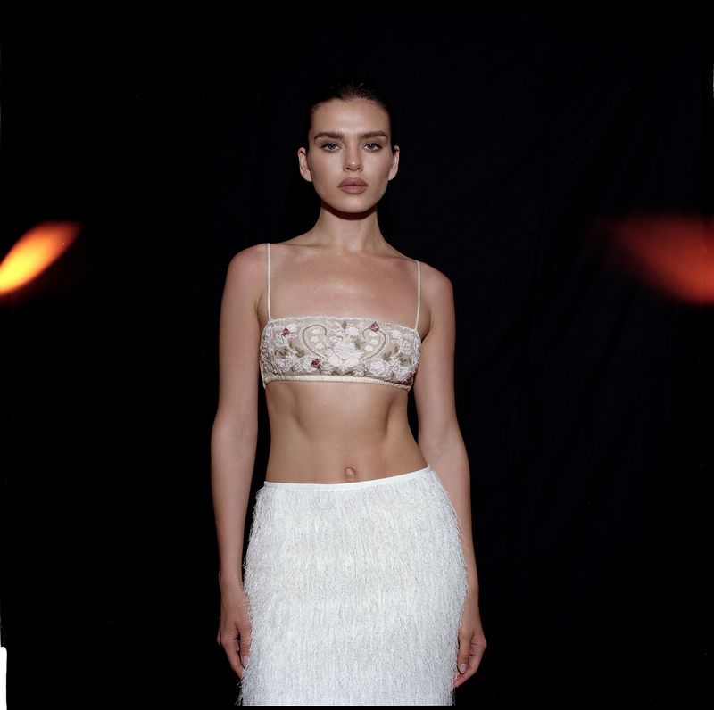Portrait of a woman wearing an embroidered strapless top and white fringe skirt against a dark background