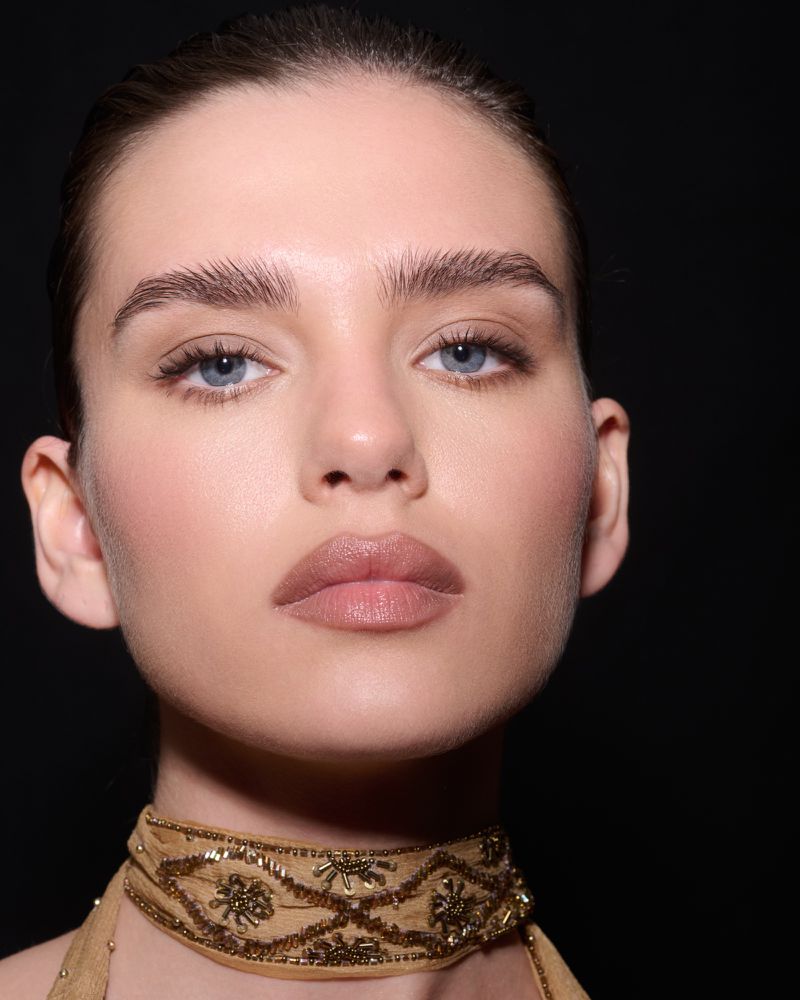 Close-up portrait of a woman with blue eyes wearing a beaded gold choker against a plain black background.