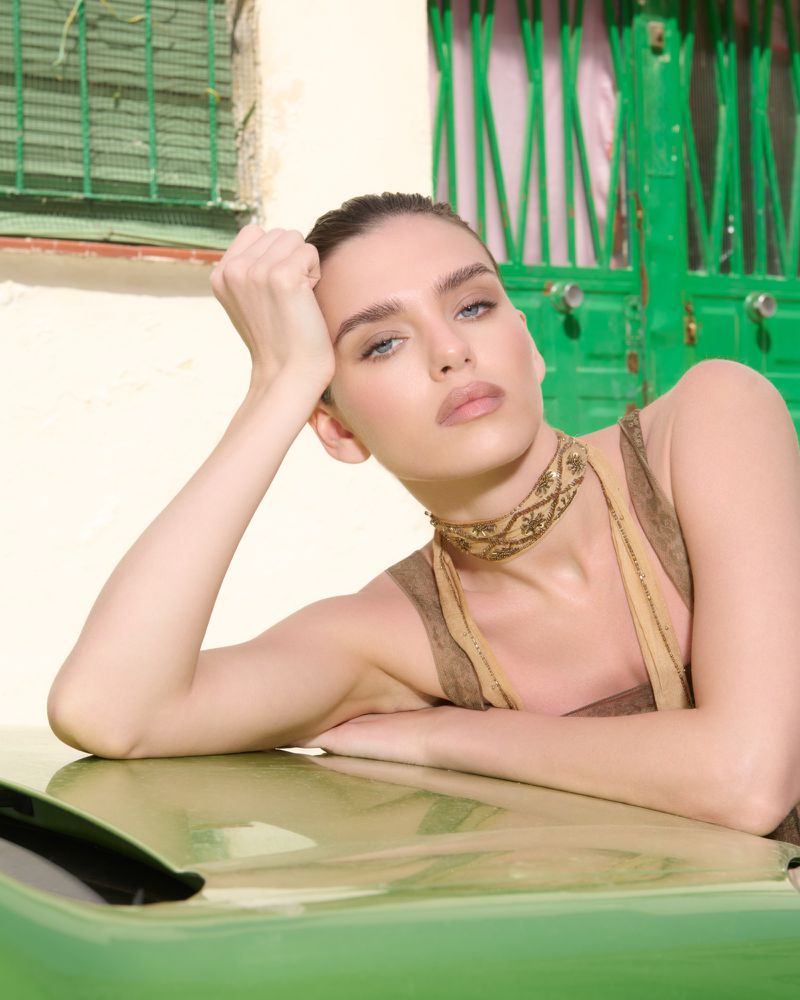 Woman resting her chin on her hand, leaning on a green vehicle with a matching green door and pale wall in the background.