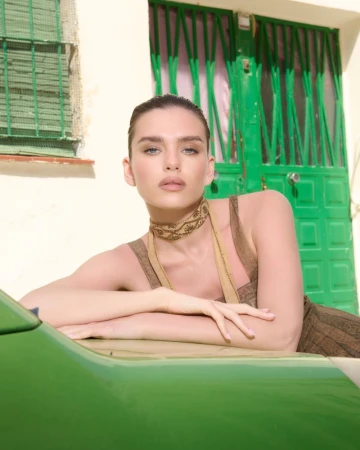 A woman in a patterned dress leaning against a green car exterior with a green doorway in the background