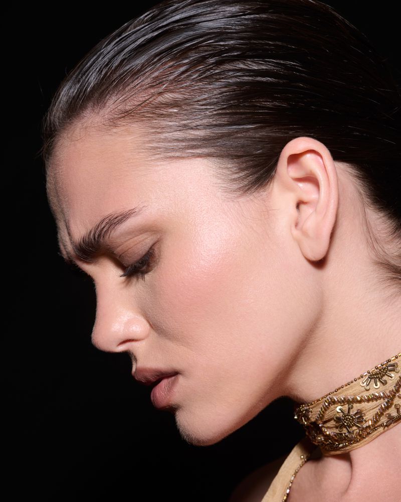 Close-up profile portrait of a woman with slicked-back dark hair wearing an embroidered gold choker against a black background.