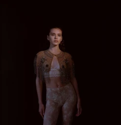 Portrait of a woman wearing a beaded cape and patterned trousers against a dark background