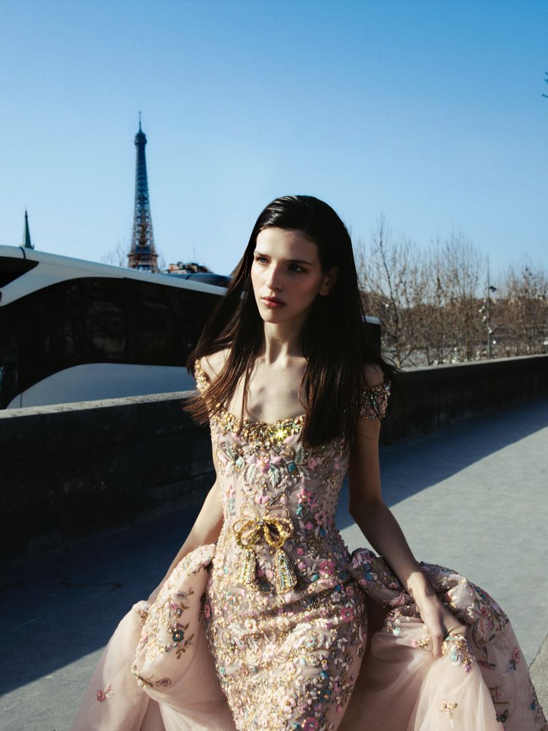 A model wearing an ornate pink and gold embroidered gown outdoors with the Eiffel Tower in the background under a clear sky.