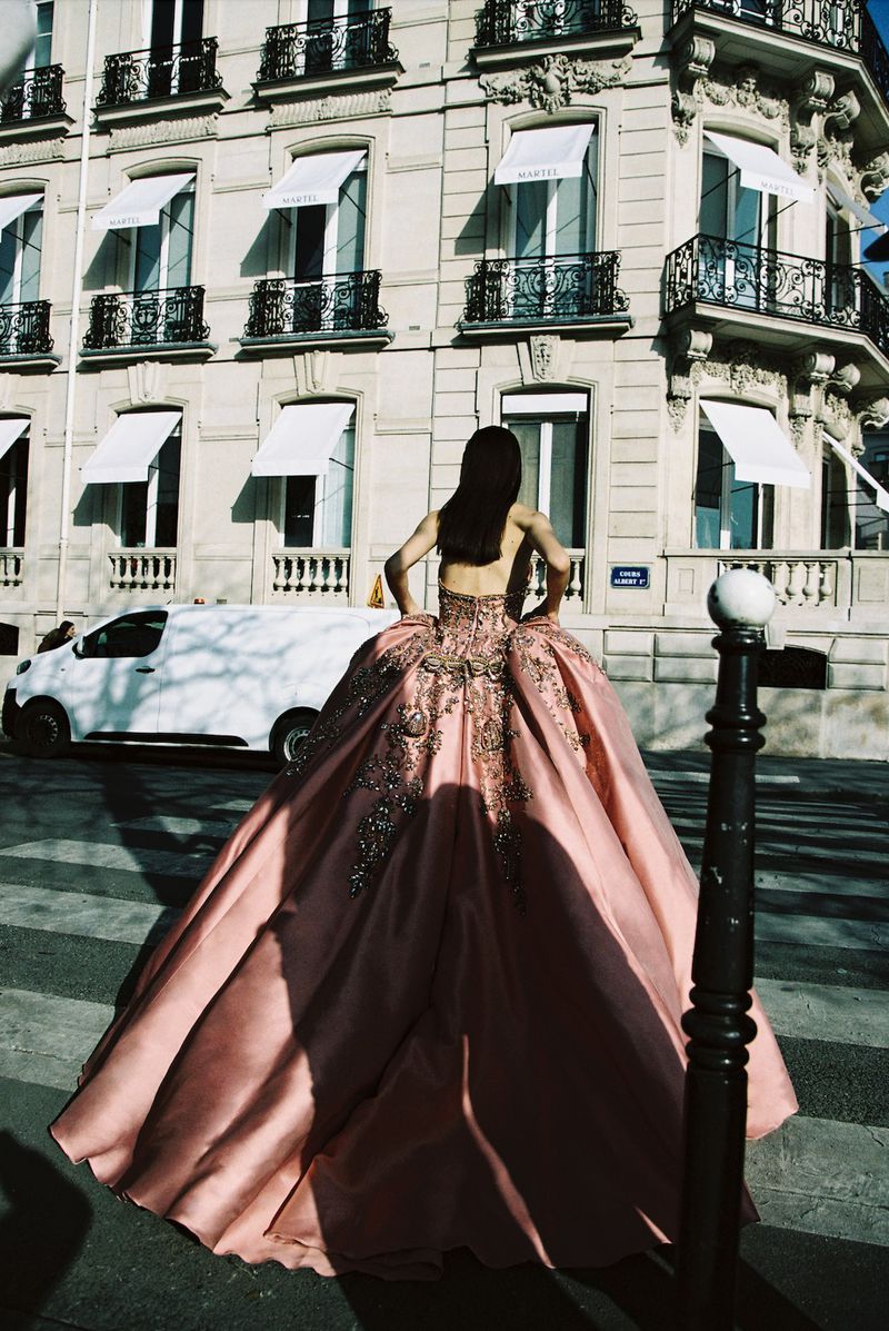Model in a voluminous pink ball gown with intricate embroidery standing on a cobblestone street in front of a classical building.