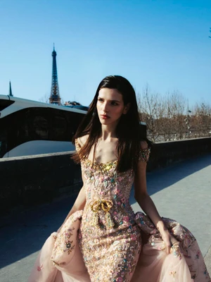 A model wearing an ornate pink and gold embroidered gown outdoors with the Eiffel Tower in the background under a clear sky.