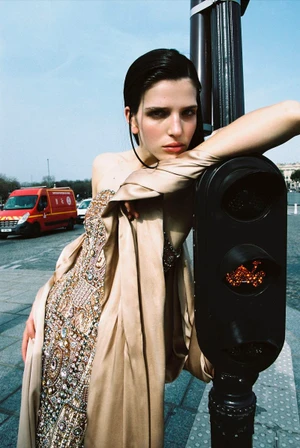 Model leaning on a traffic light pole wearing an embellished beige gown against an urban street background.