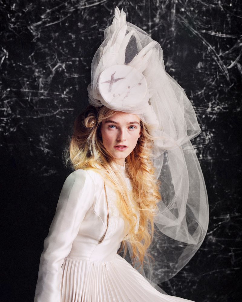 Model wearing a white dress and elaborate feathered tulle hat against a dark marbled background