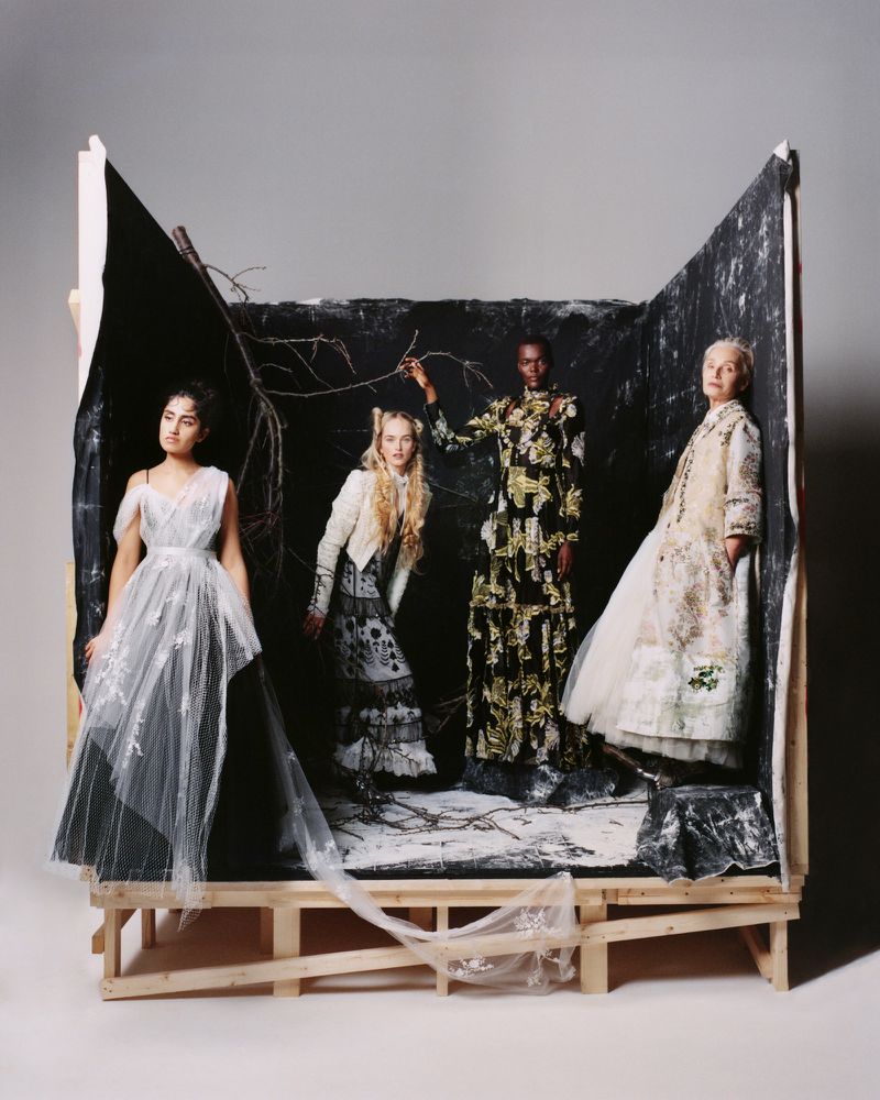 Four women in patterned and tulle gowns standing inside a rustic wooden diorama set on a white floor for British Vogue