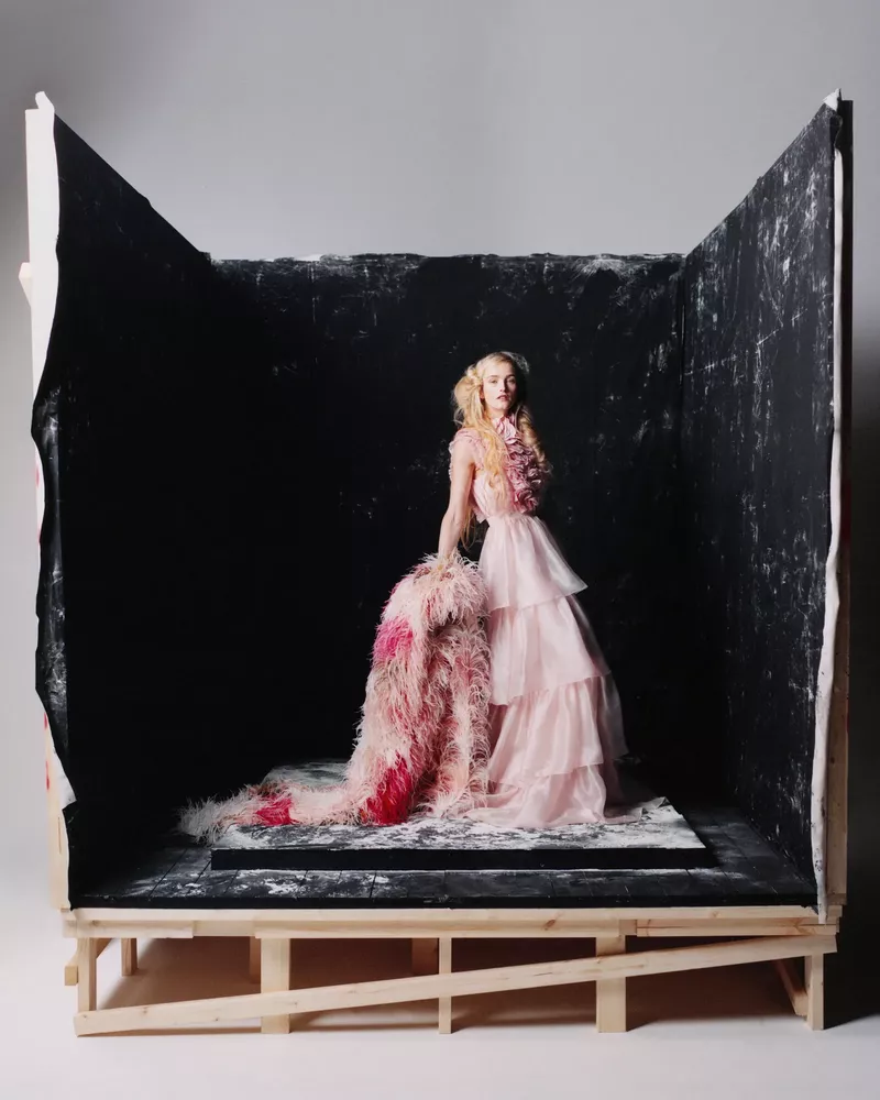 A fair-skinned model with long blonde hair poses in a tiered pink dress inside a black and white painted wooden box.