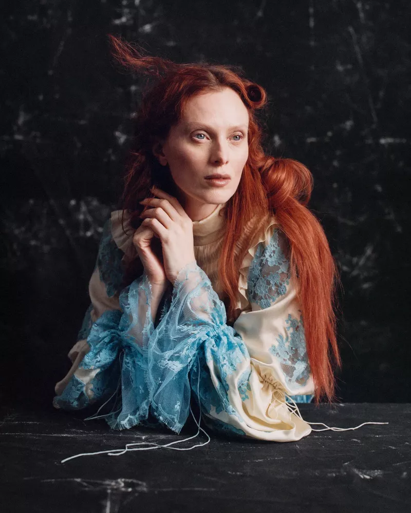 Model Karen Elson with long red hair wearing a patterned dress leaning against a dark textured surface for British Vogue.