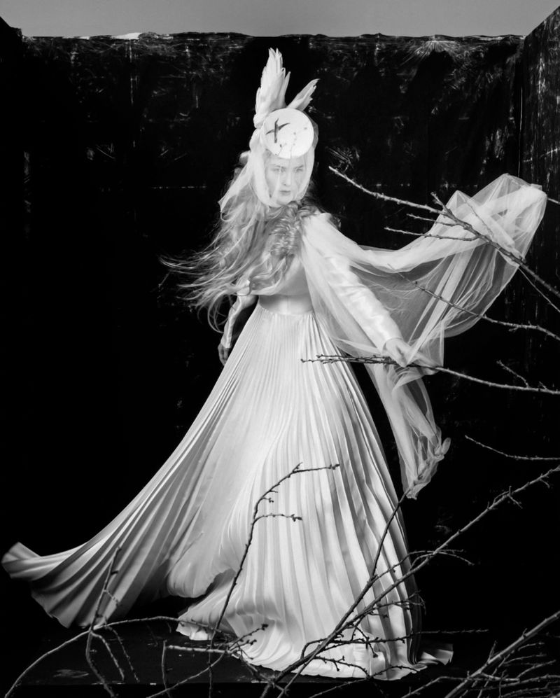 Model in a white pleated gown and sheer feathered headpiece standing among dark branches in a studio setting