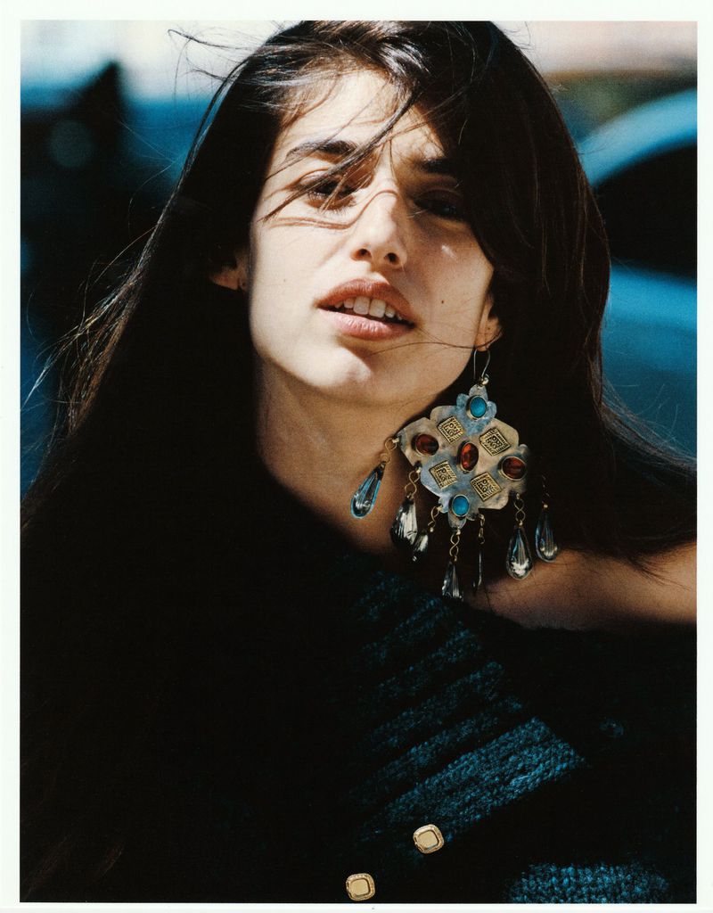 Portrait of a woman wearing an ornate metal earring with blue stones and a dark textured top outdoors in natural light
