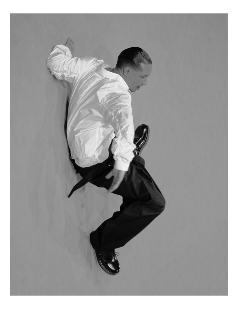 Monochrome studio shot of a man in a white shirt and dark trousers, leaning dynamically against a grey wall, showcasing movement and style.