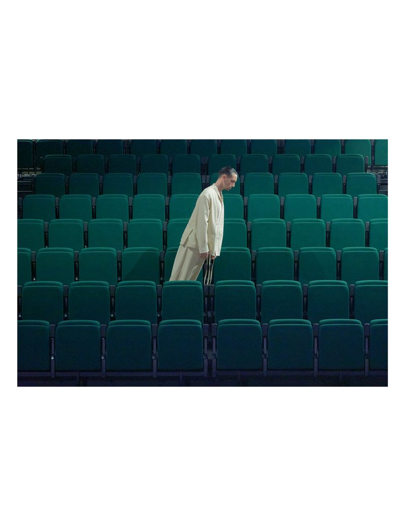 A man in a cream suit standing amidst rows of empty teal auditorium seats for Dapper Dan Magazine editorial.