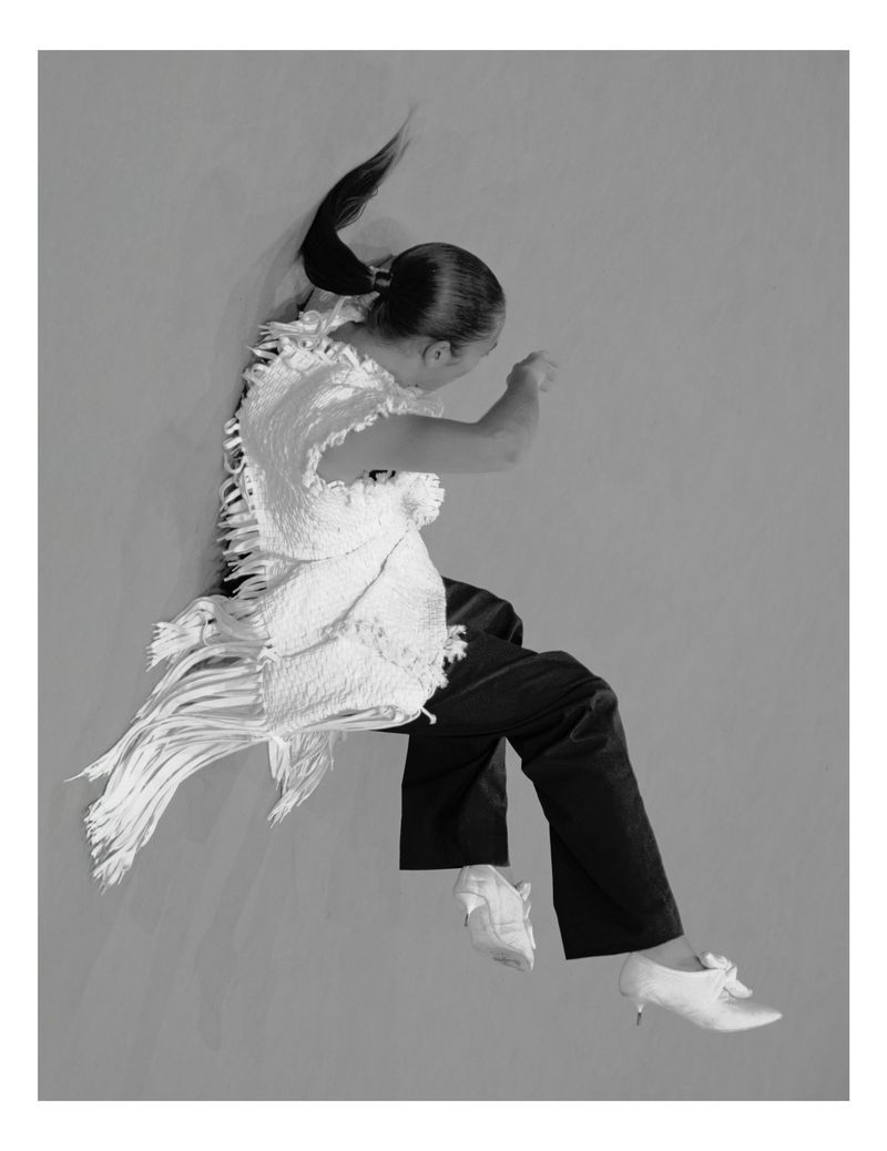 Model in white fringed top and dark trousers captured mid-jump against a grey seamless background