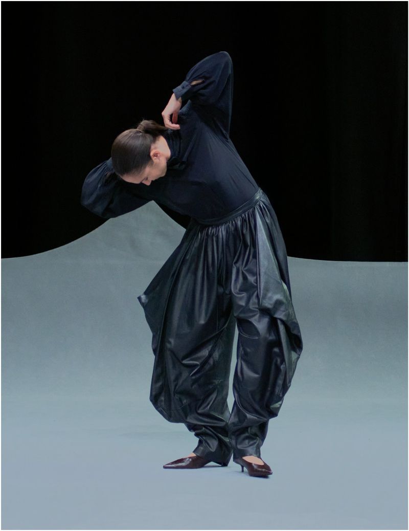 A model in a black long-sleeve shirt and dark trousers poses gracefully against a grey studio backdrop.