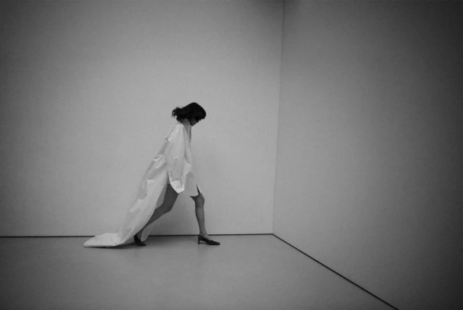Model wearing an oversized white shirt with a long train walking across a minimal corner studio space