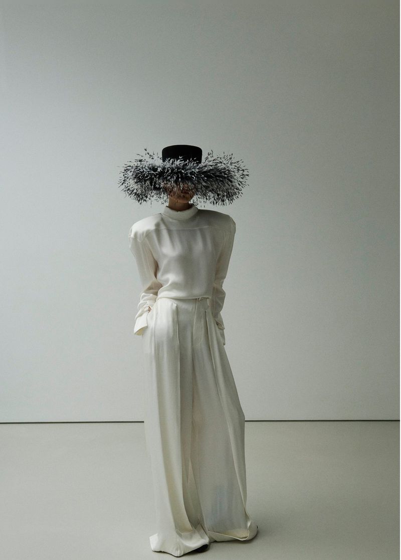 A person wearing a cream-colored outfit and a large black hat with dark fringe, standing against a plain white background in a studio setting.