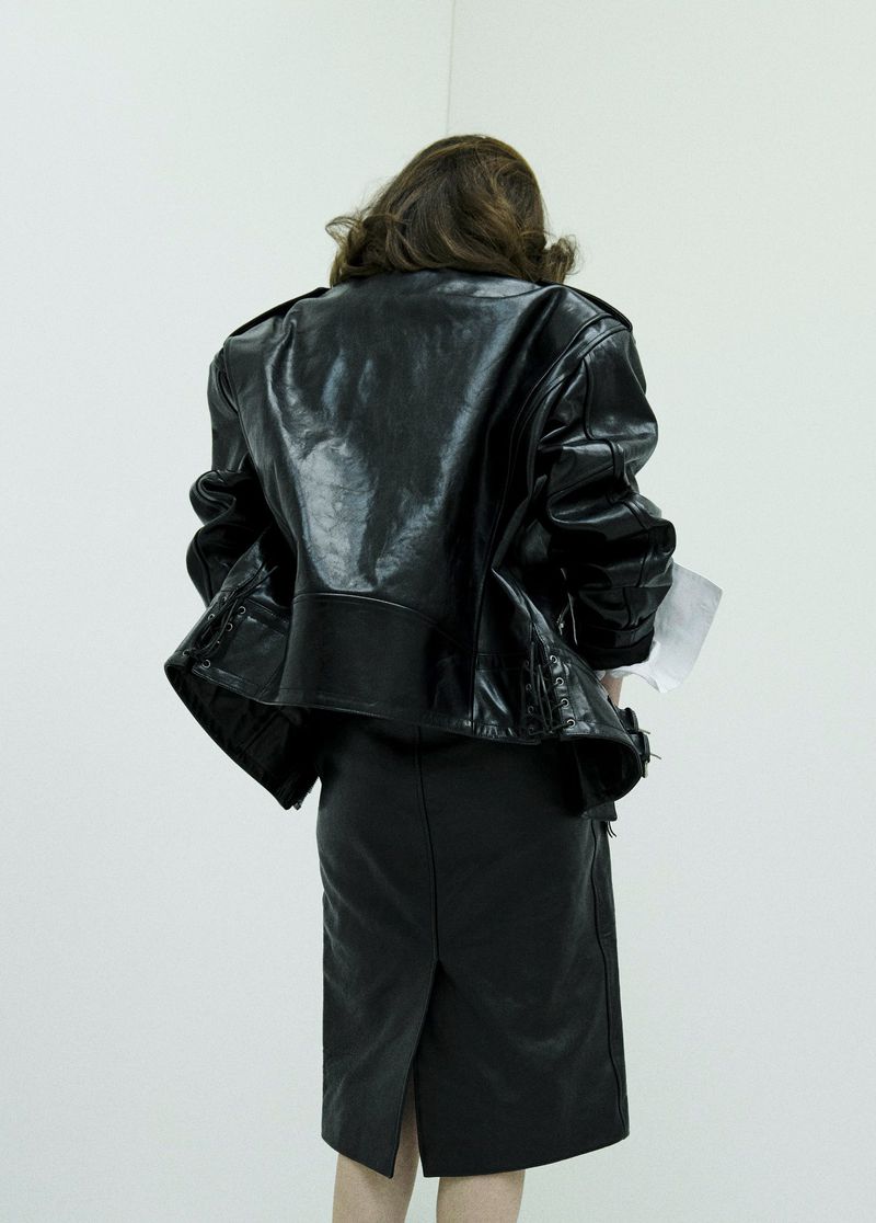 Model from behind wearing a black leather jacket and skirt against a plain white studio wall for Puss Puss Magazine