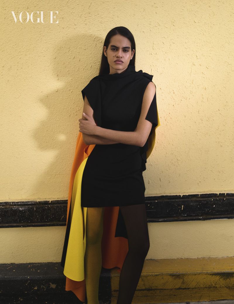 Model in a black dress with a yellow and orange cape standing against a textured yellow wall for Vogue Arabia.