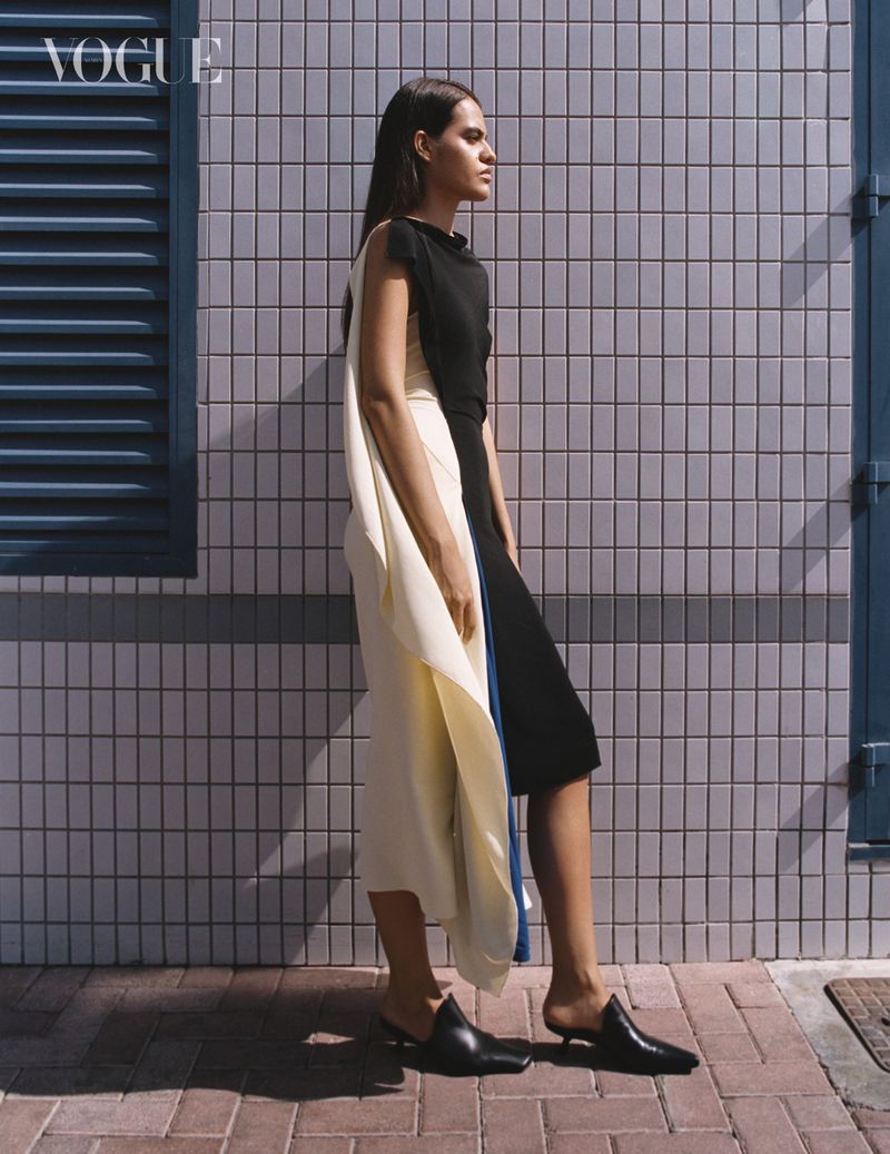 Woman wearing a black and cream asymmetrical dress standing against a tiled exterior wall for Vogue Arabia.