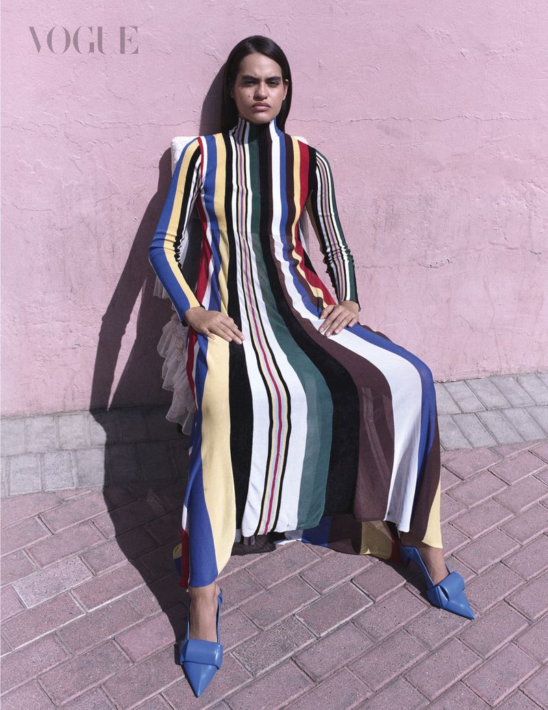 Model seated on a chair against a pink wall wearing a colorful striped turtleneck dress and blue heels