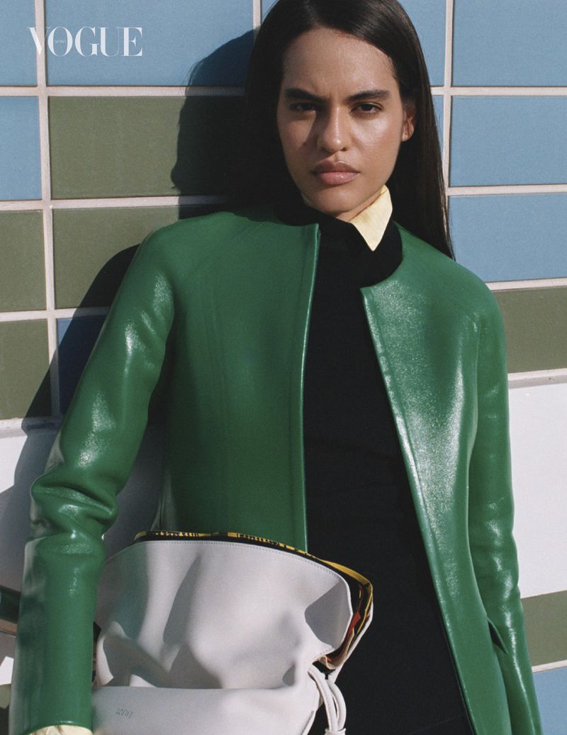 Model wearing a green jacket and holding a white handbag against blue and green tiled walls for Vogue Arabia.