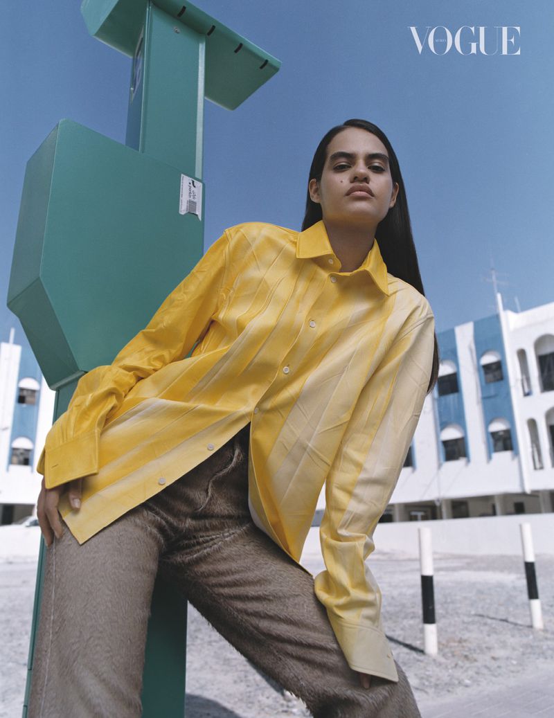 Model in a yellow button-down shirt and brown textured pants leaning against a green structure under a blue sky.