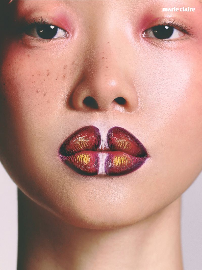 Close-up portrait of a woman with avant-garde pink eyeshadow, subtle freckles, and intricate metallic red and gold lip art for Marie Claire Colombia.