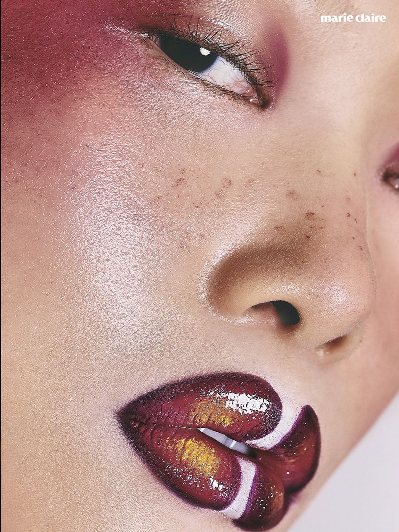 Close-up beauty portrait of a person with dramatic pink eyeshadow, freckles, and glossy dark red and gold metallic lips.